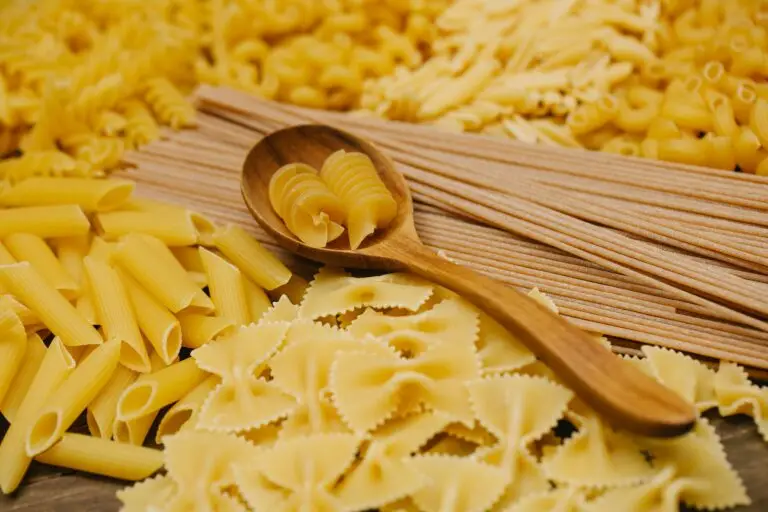 A selection of uncooked Italian pasta, including penne, farfalle, and rotini, on a wooden spoon.