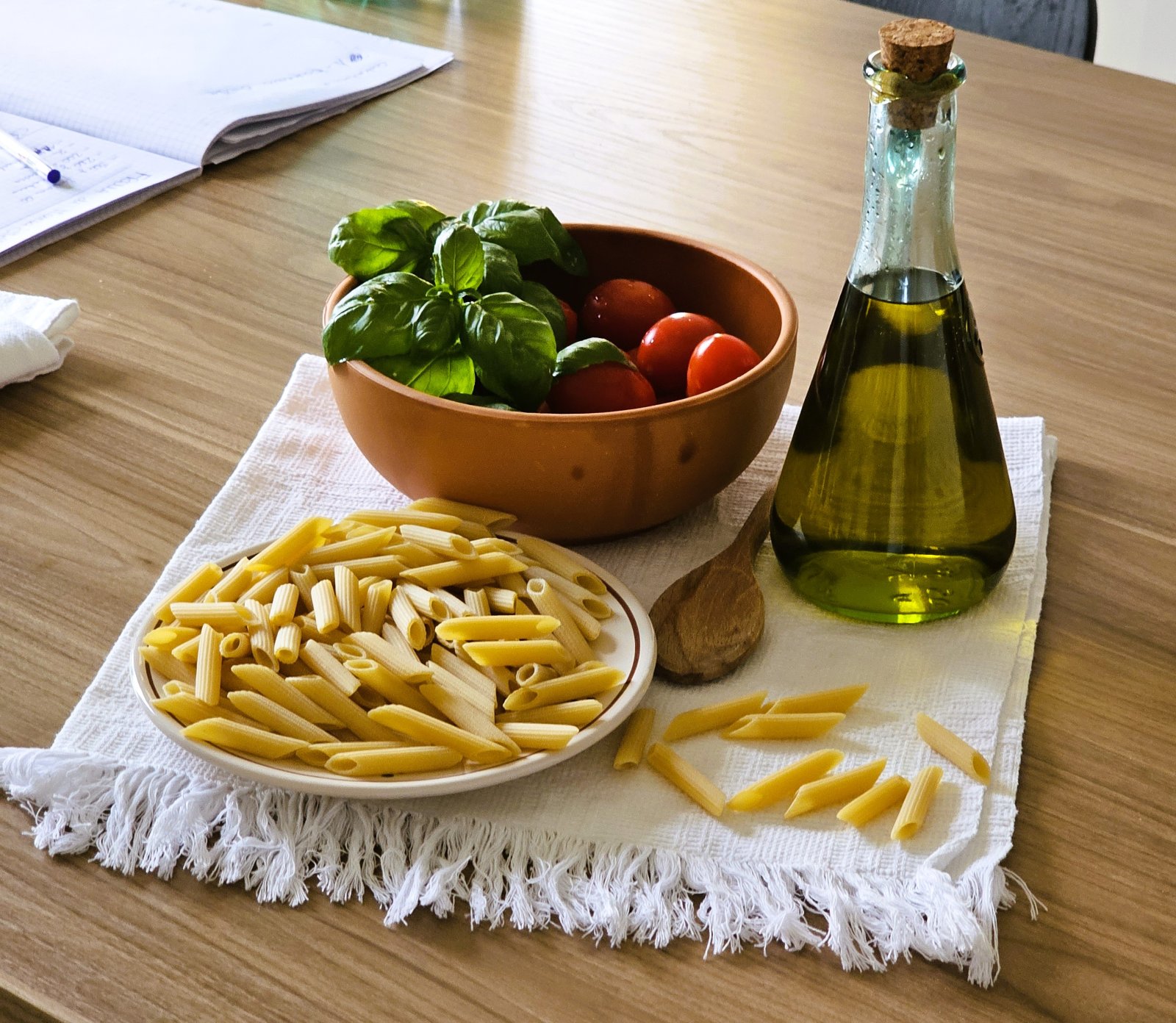 Pasta, pomodori, basilico e olio extravergine su un tavolo da cucina con quaderno di appunti.